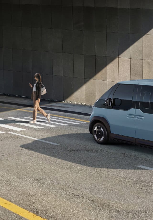 Der Kia PV5 Passenger hält am Zebrastreifen, während ein Fußgänger vorbeigeht.